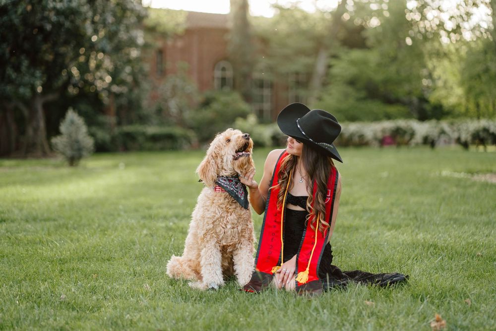Woman in graduation cap and red gown sits on grass with a golden doodle dog wearing a bandana.