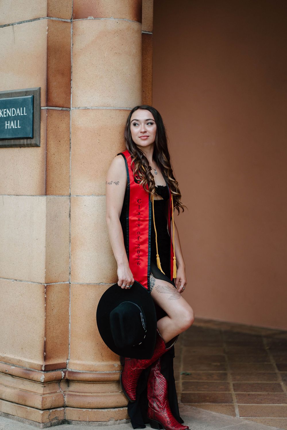 Young woman in red graduation stole and cowboy boots holds black hat beside Kendall Hall column.