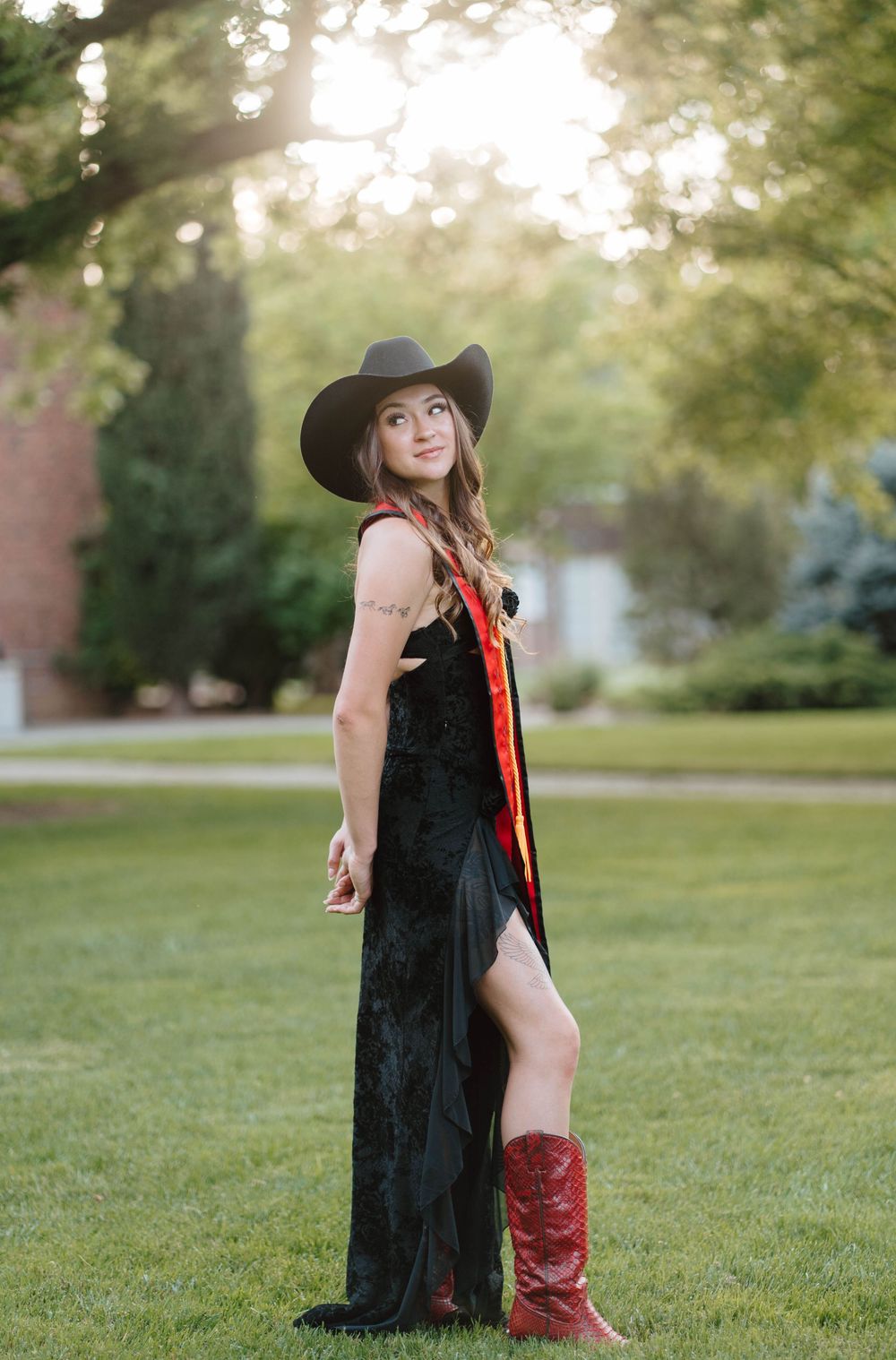 Young woman in black velvet dress, cowboy hat, and red boots poses outdoors on green grass at golden hour.