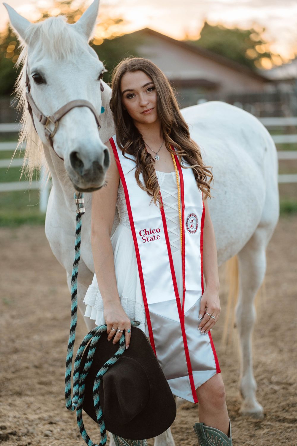 Chico State graduate in white dress and red sash poses with white horse holding cowboy hat at golden hour.
