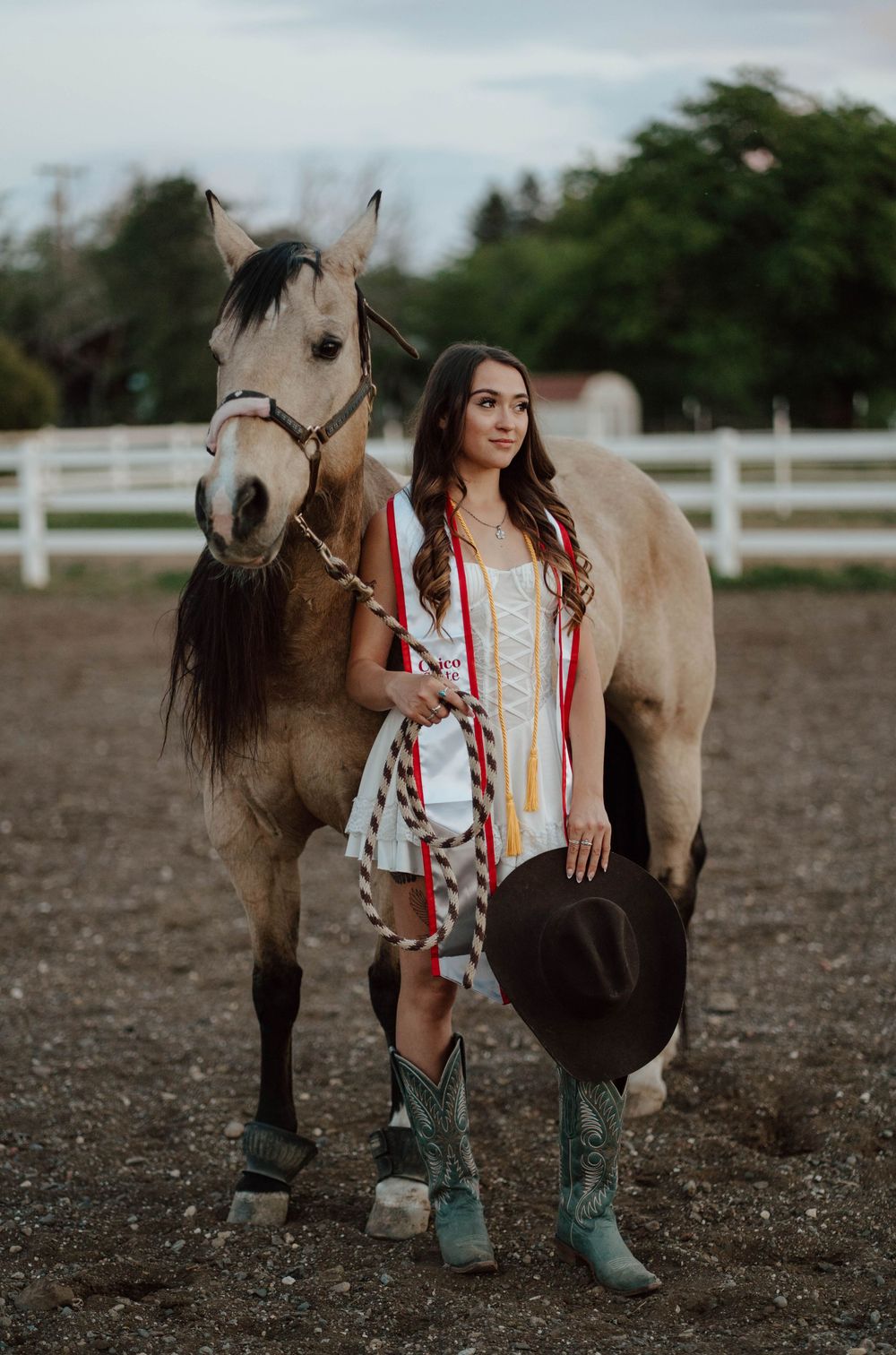 A young woman in a white dress and teal cowboy boots holds a hat beside a buckskin horse at a ranch.