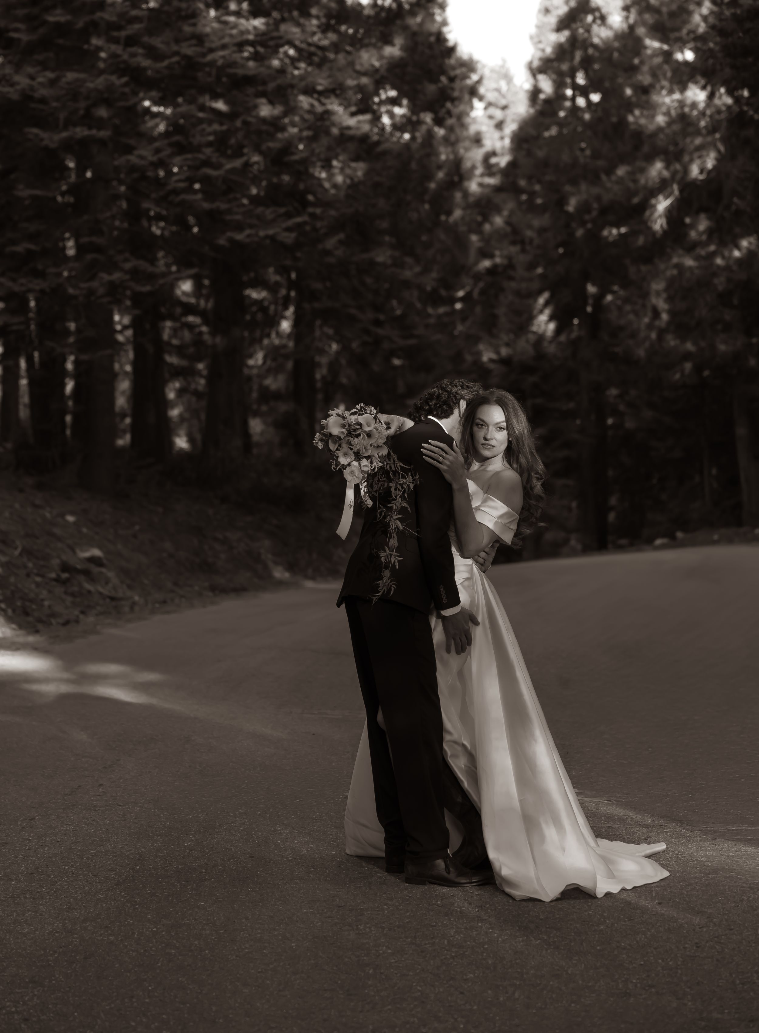 Black and white photo of a bride and groom embracing on a road surrounded by tall trees, bride holding a bouquet.