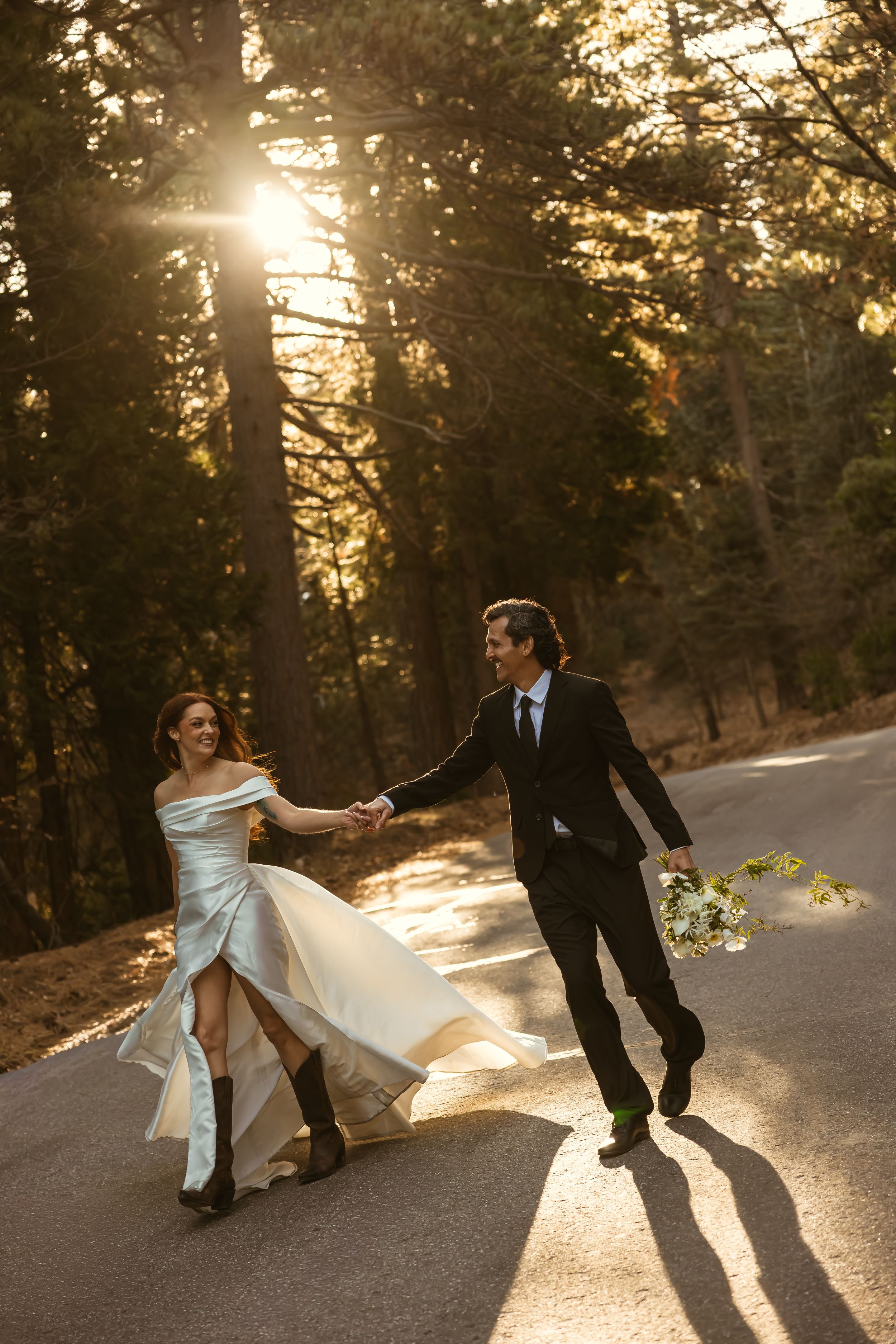 Bride and groom walking hand-in-hand down a sunlit forest road, bride holding her flowing white gown.