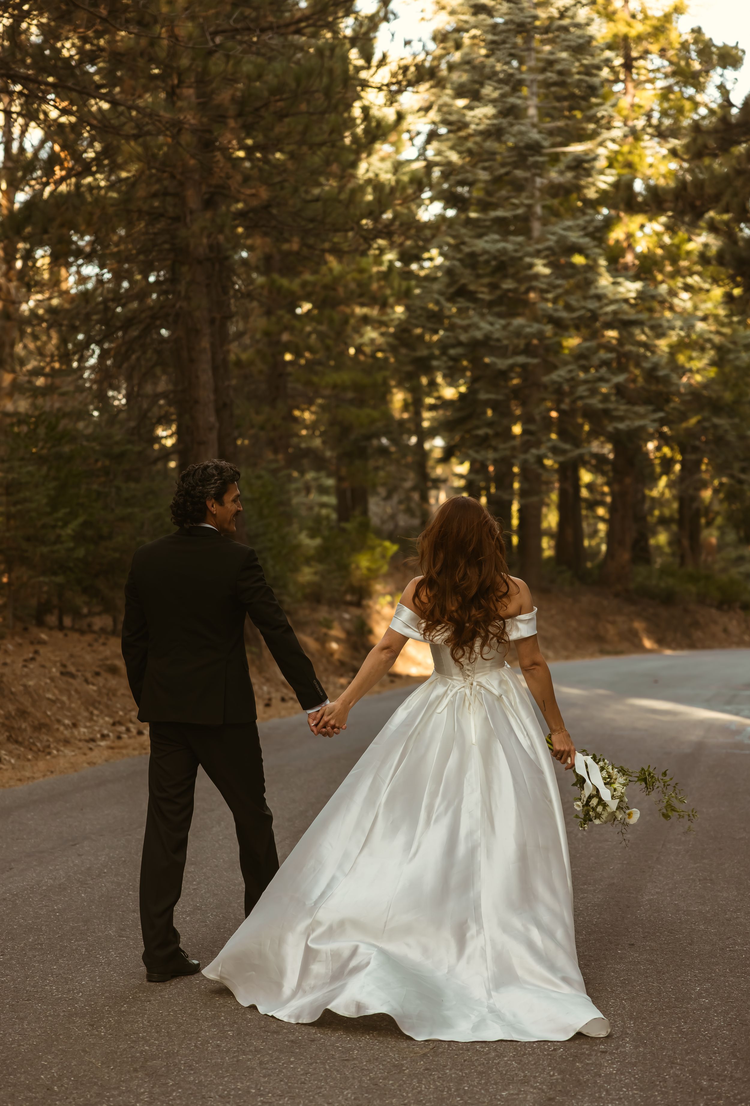 Bride in a white off-shoulder gown holds hands with a groom in black suit on a forest road at golden hour, Los Angeles Wedding Photographer