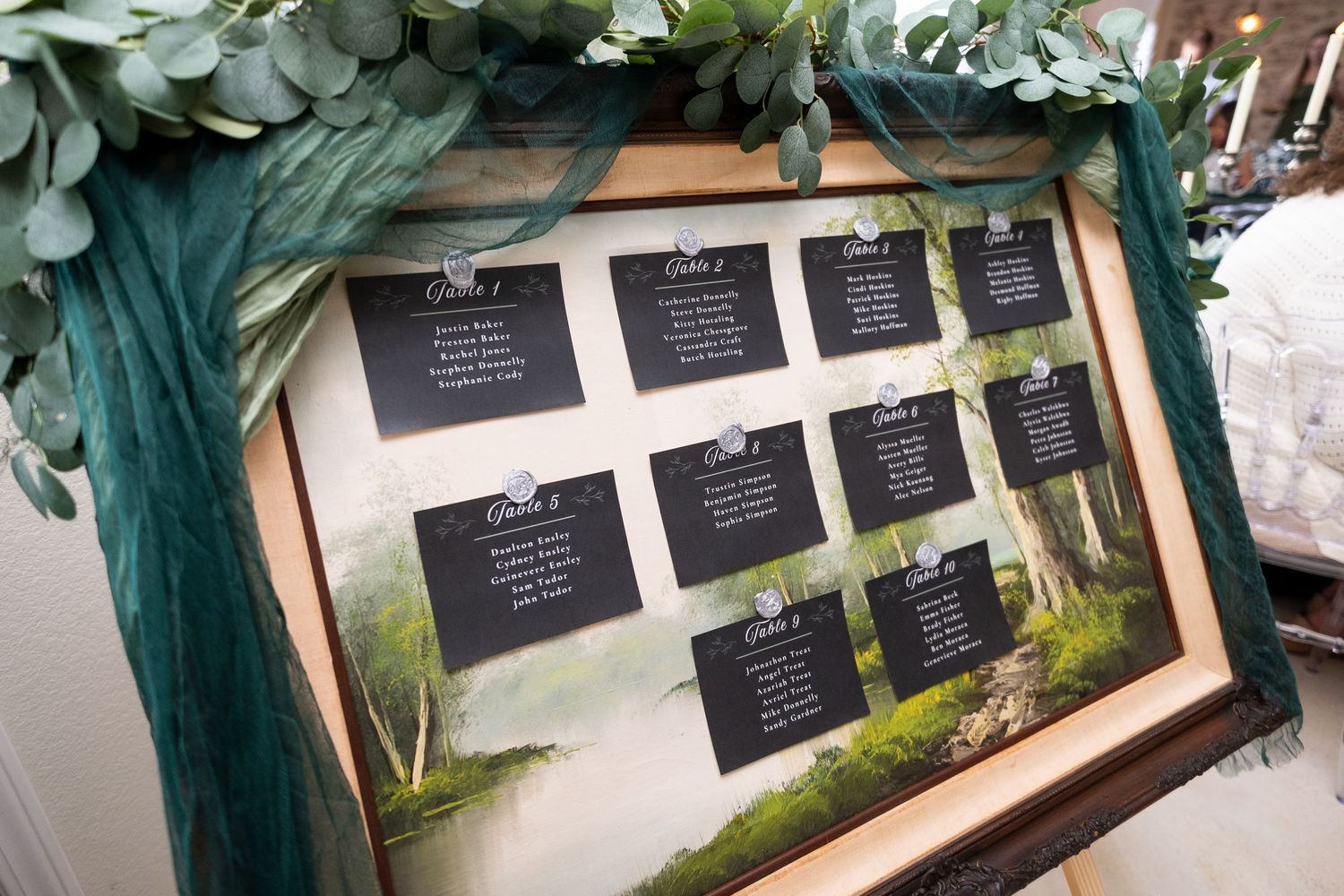 Elegant wedding seating chart display in a wooden frame with dark cards and greenery garland decoration.