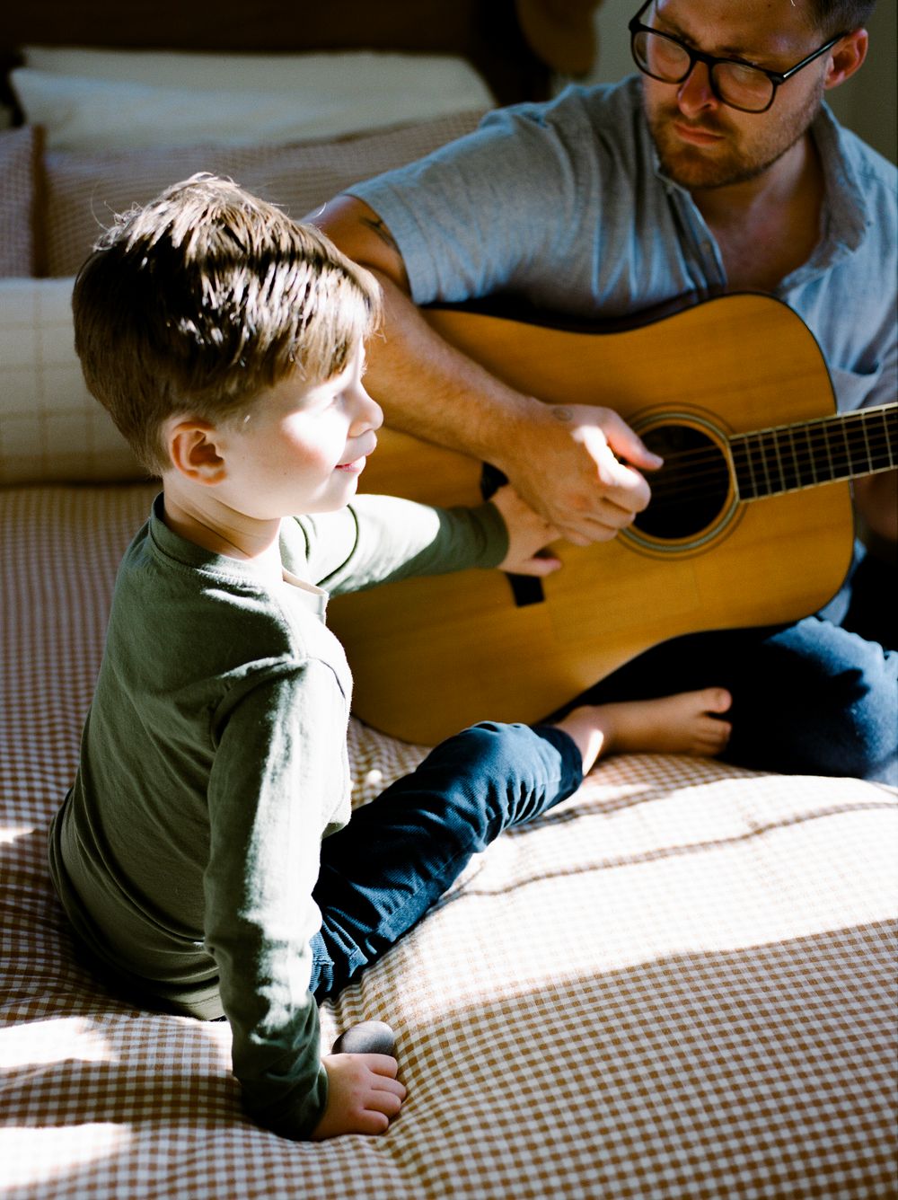A father plays acoustic guitar for his young son while sitting together on a bed in warm natural light.