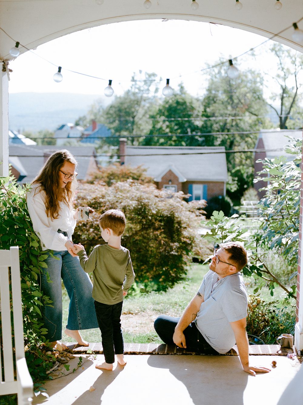 Family of three enjoying a sunny porch, with mom, dad, and young child playing outdoors with lush garden views.