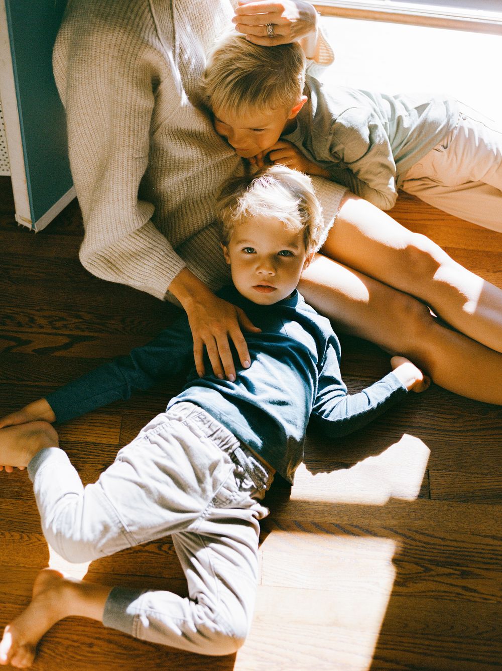 Two young children sitting on hardwood floor in natural light, older sibling hugging younger toddler in casual home setting.