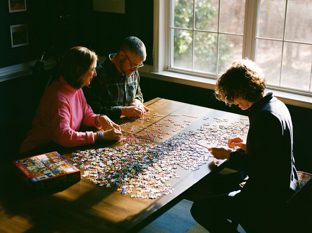 Three people working together on a large jigsaw puzzle spread across a wooden table near a sunlit window.