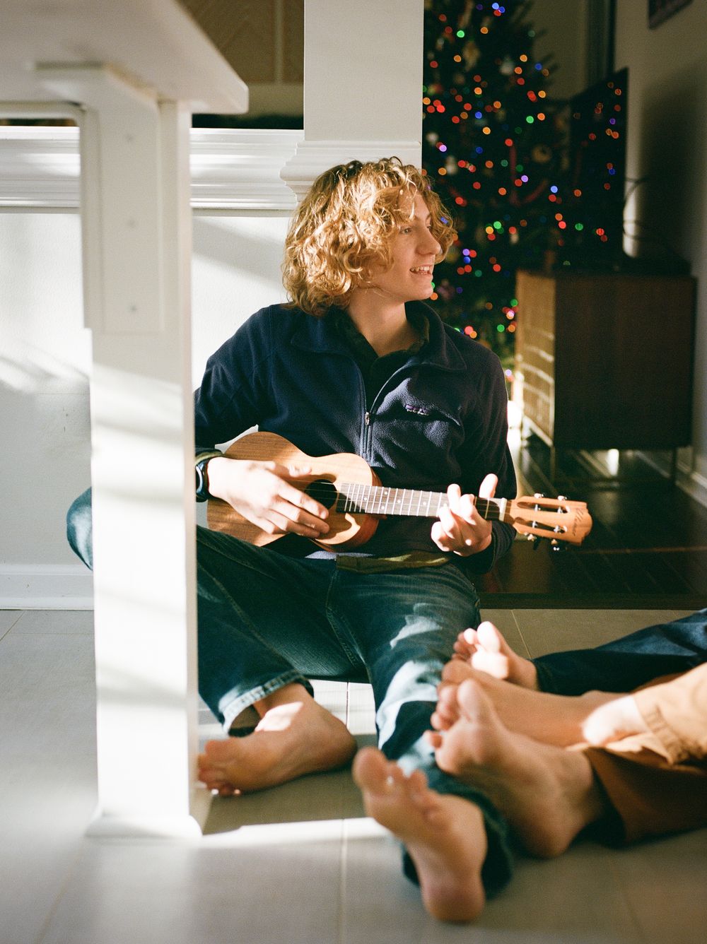 Person with curly hair playing ukulele barefoot indoors near a decorated Christmas tree in warm sunlight.