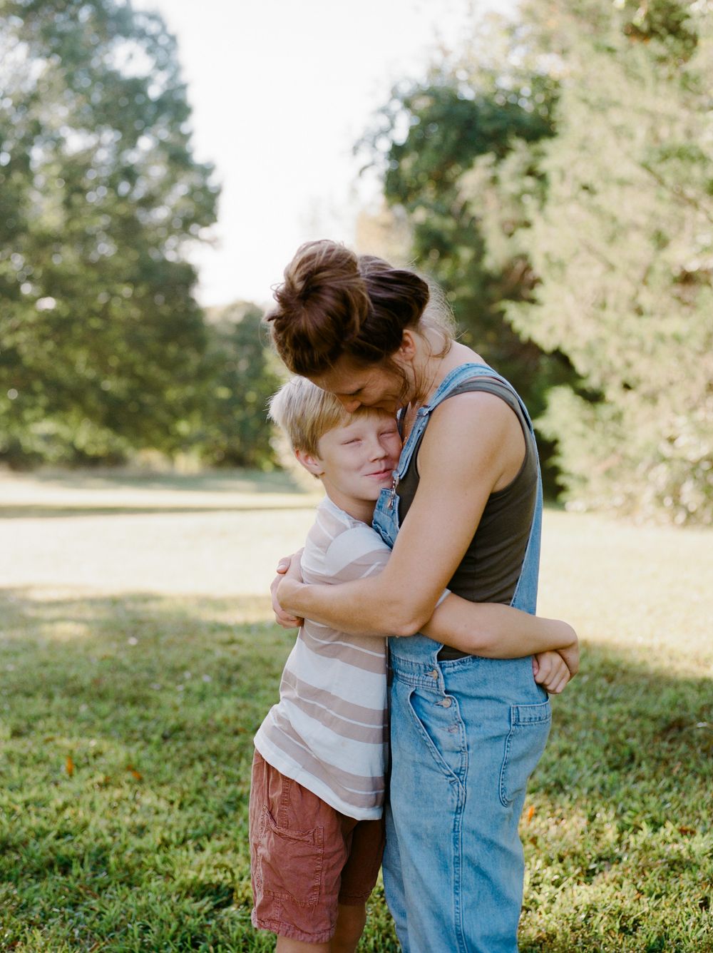 A mother tenderly hugs her young blonde son outdoors in a lush green park on a sunny day.