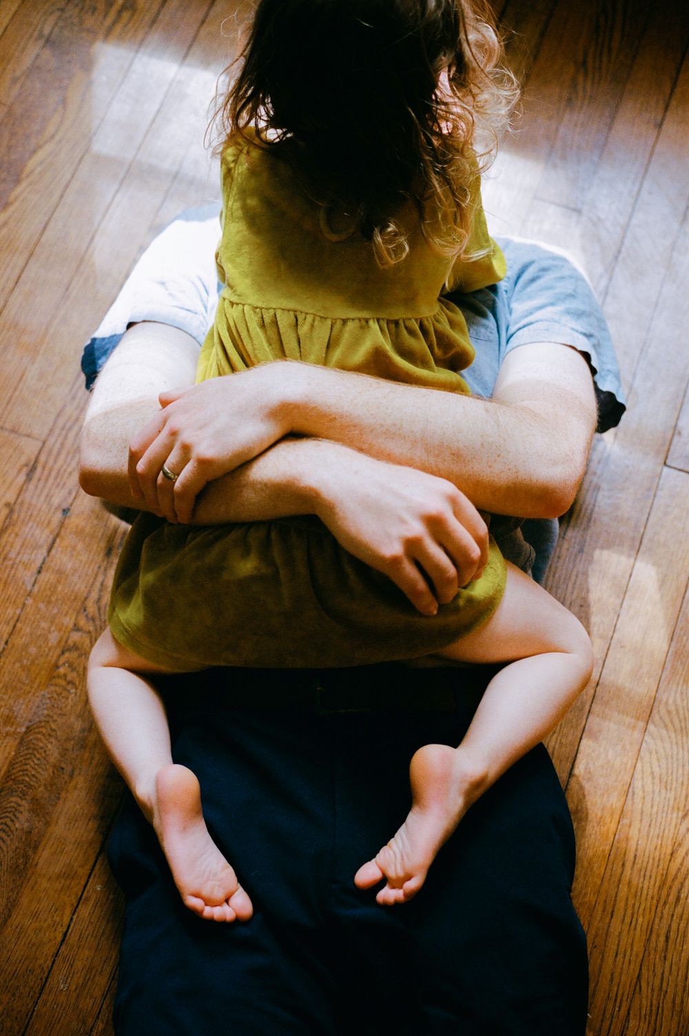 Adult sitting on wooden floor holding a young child in a yellow dress between their legs.