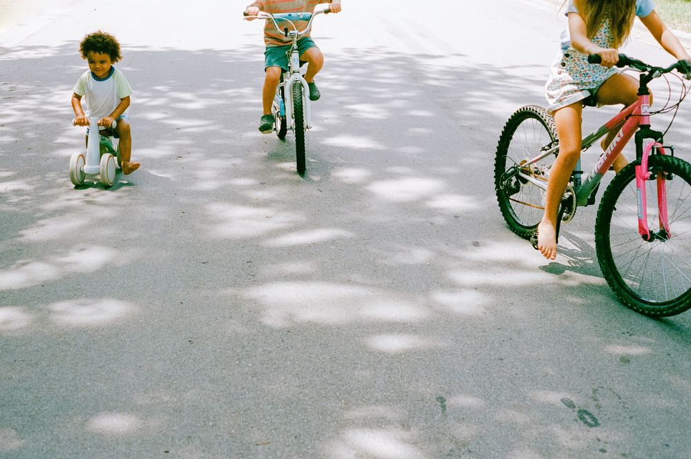 Three children playing outdoors, two riding bicycles and one on a scooter, on a sunlit pavement with dappled shadows.