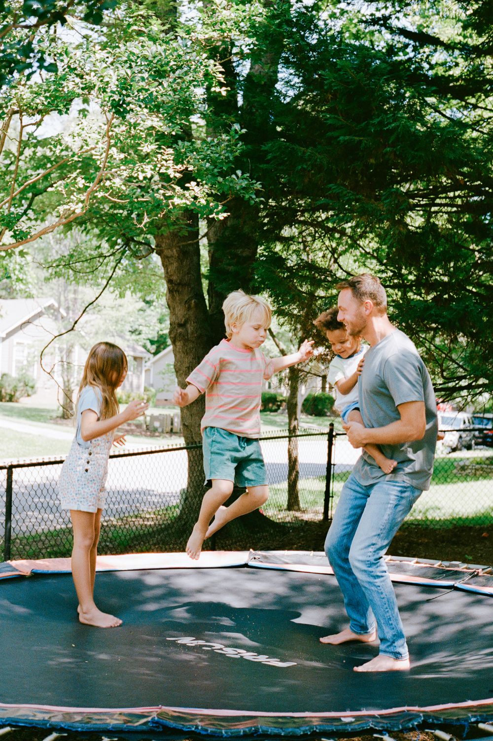 A father and three children joyfully jumping on a backyard trampoline surrounded by lush green trees on a sunny day.