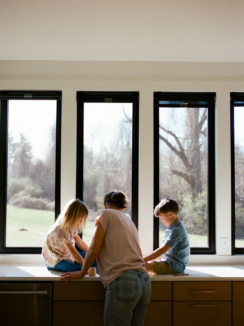A parent and two children sit on a kitchen counter looking out large black-framed windows at a green yard.
