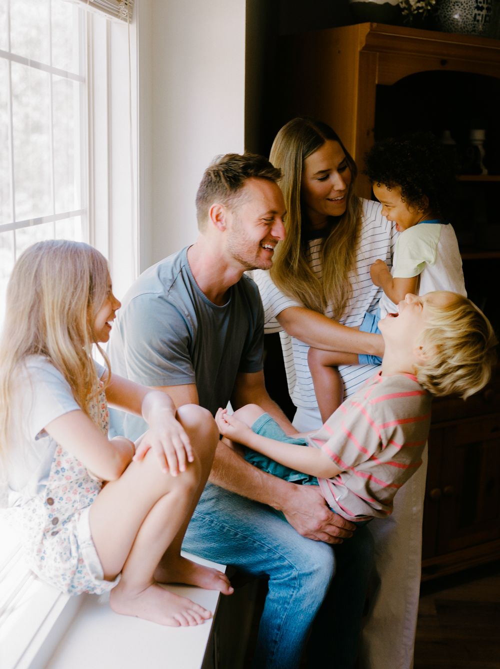 Happy family of five laughing together by a bright window in a cozy home setting.