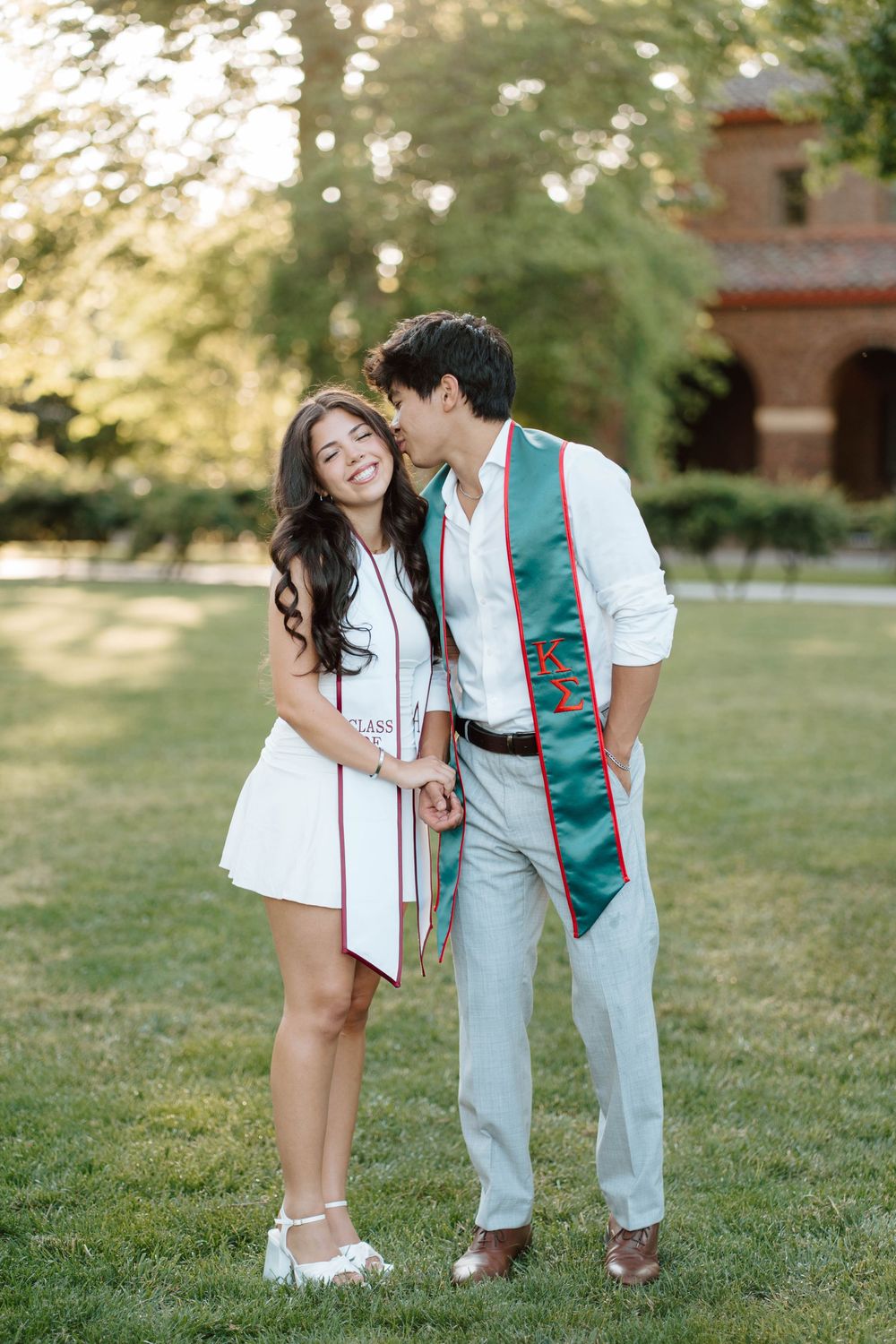 A couple in graduation stoles celebrate outdoors on a green campus lawn, sharing a tender kiss moment.