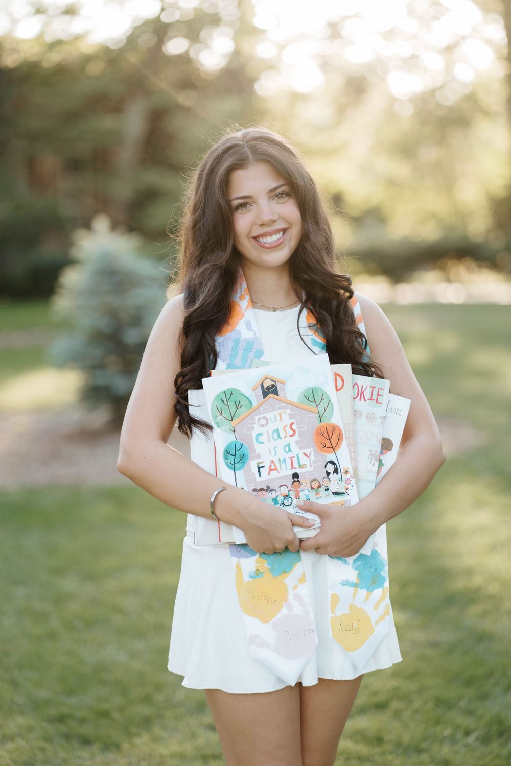Smiling young woman in floral romper holding colorful children's books outdoors in a sunlit garden setting.