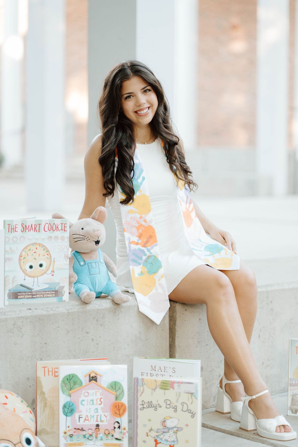 Smiling woman in white dress sits among children's books including The Smart Cookie and Mae's First Day.