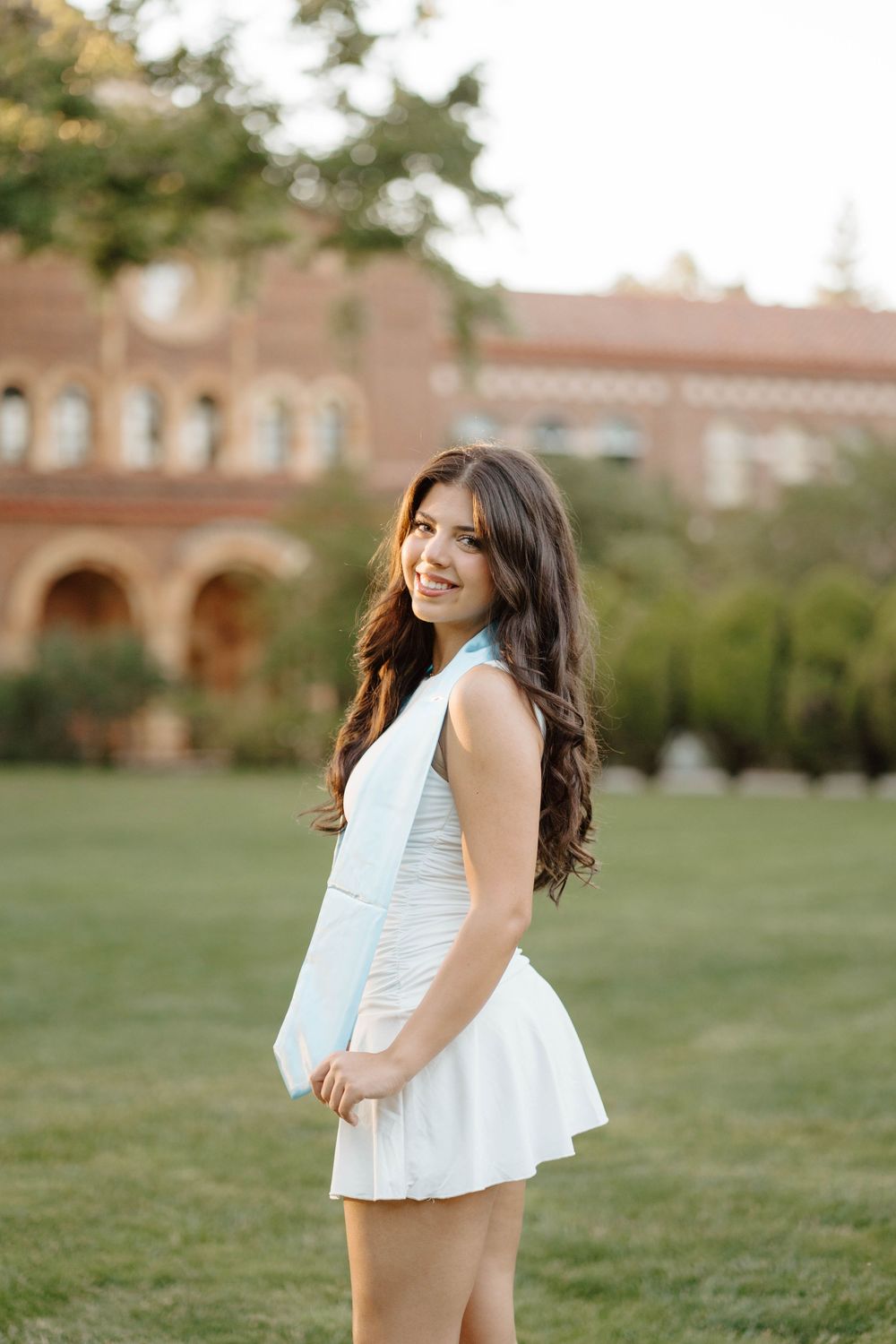 Young woman in white dress smiling outdoors on green lawn near brick building.