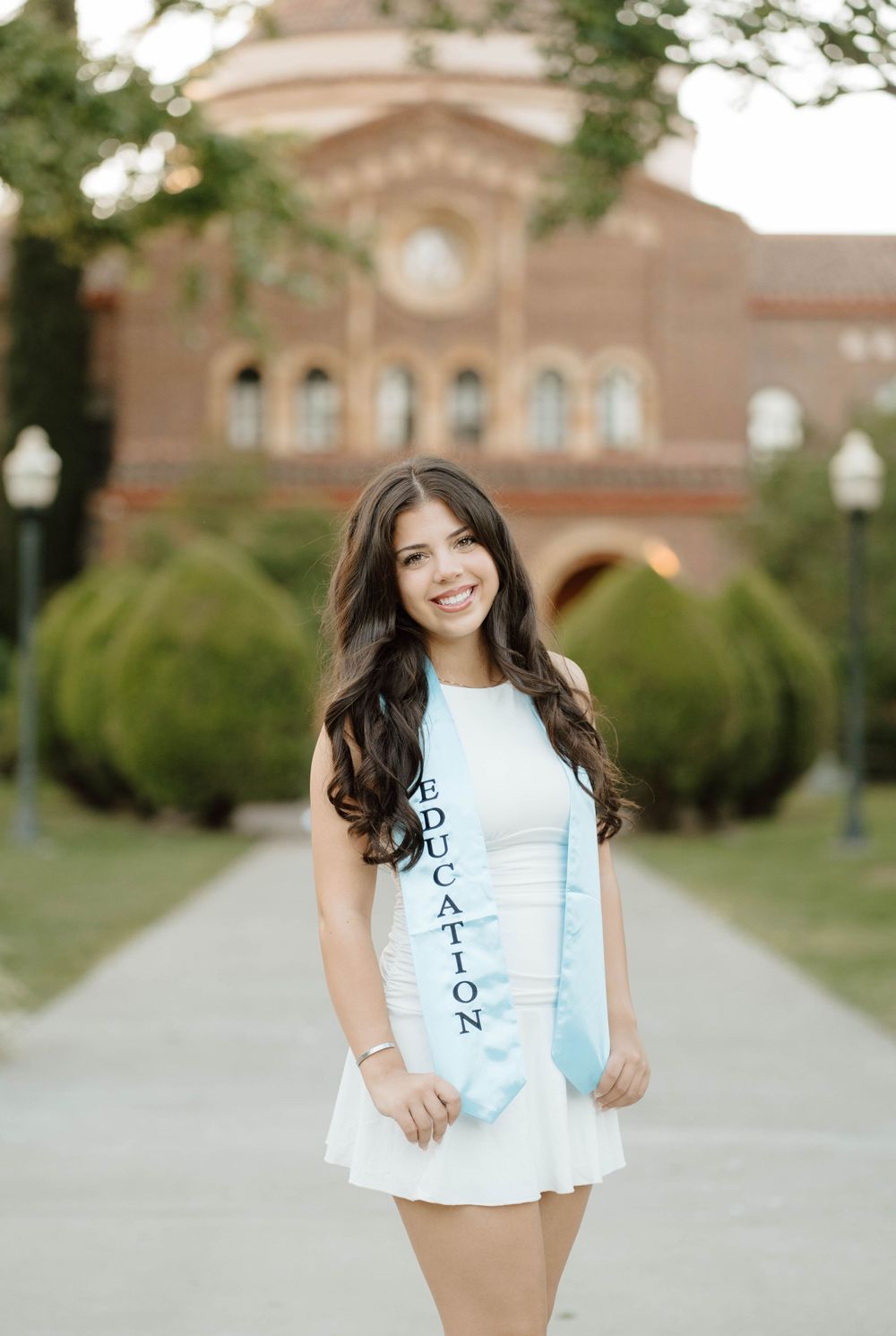 Young woman in white dress wearing an Education sash poses outdoors in front of a red brick university building.