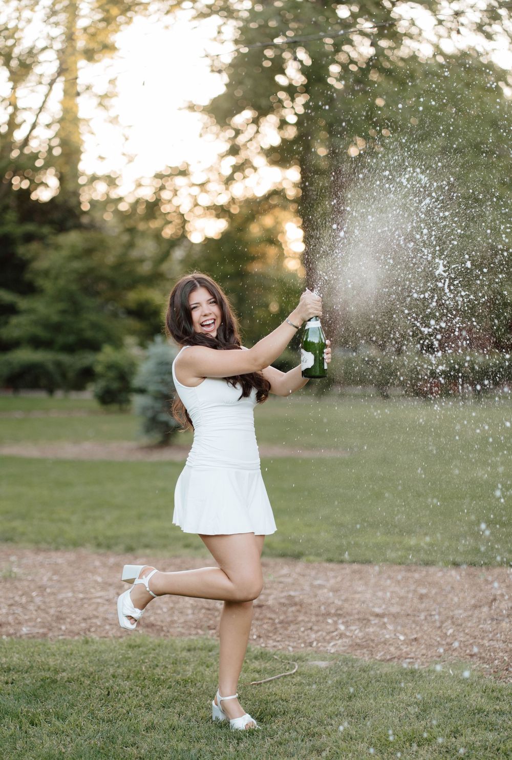 Young woman in white dress joyfully popping champagne bottle outdoors in a park at golden hour.
