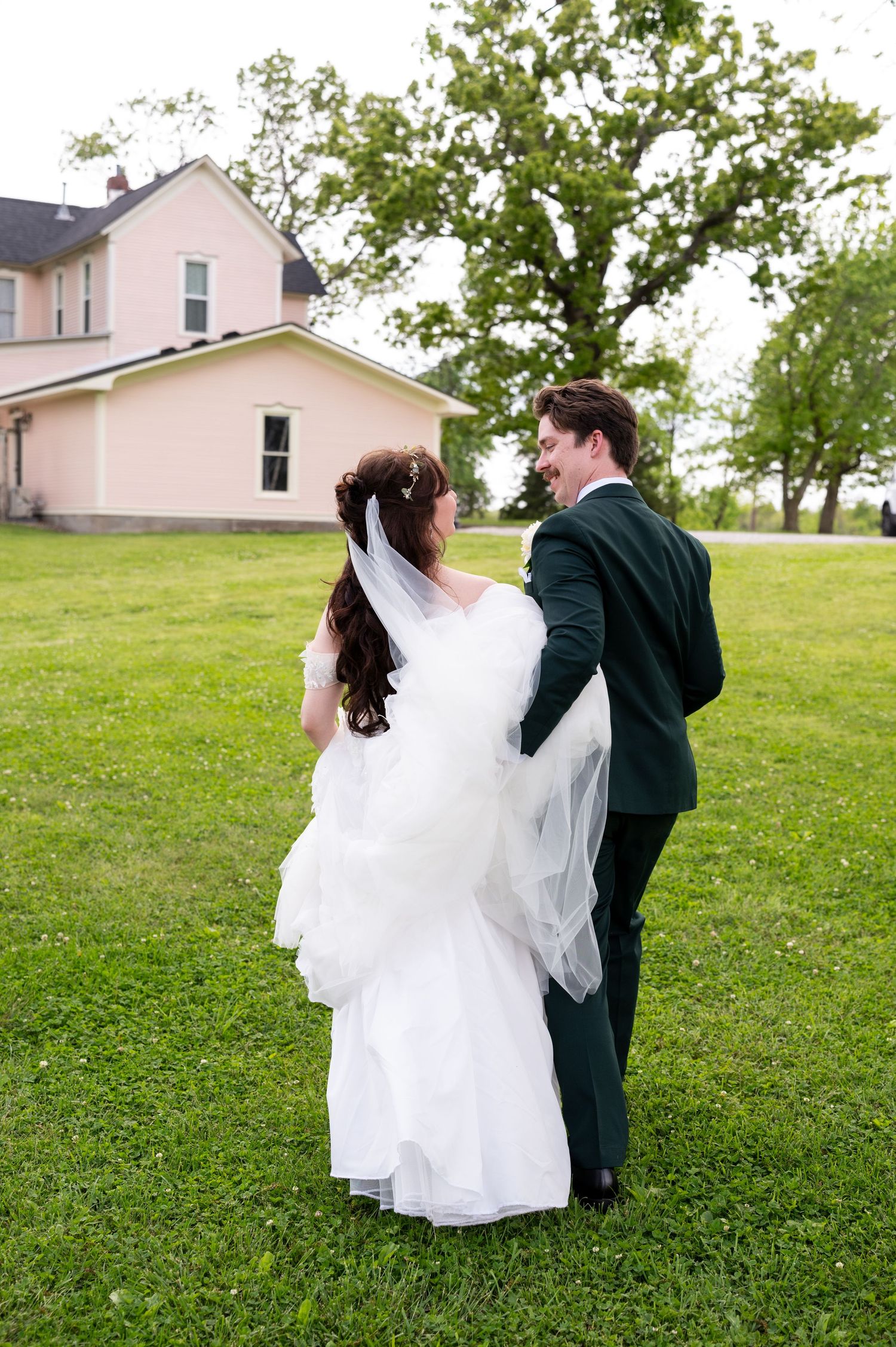 Bride in white gown and groom in dark green suit share a joyful moment on green lawn near a pink farmhouse.