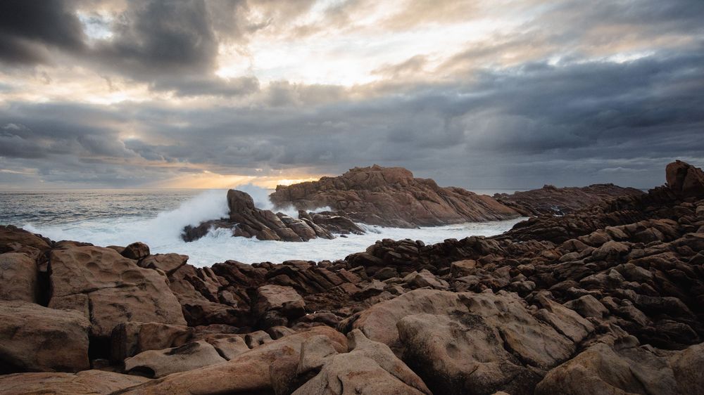 Rocky coastline with crashing waves under dramatic storm clouds at sunset, showcasing rugged coastal landscape.