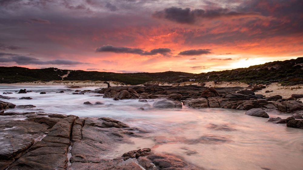 Rocky coastal shoreline at dramatic sunset with vivid red and orange sky reflecting on misty ocean waves.