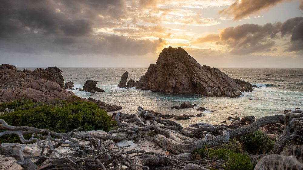 Rocky coastal landscape at sunset with dramatic clouds, ocean waves, and driftwood in the foreground near rugged sea stacks.