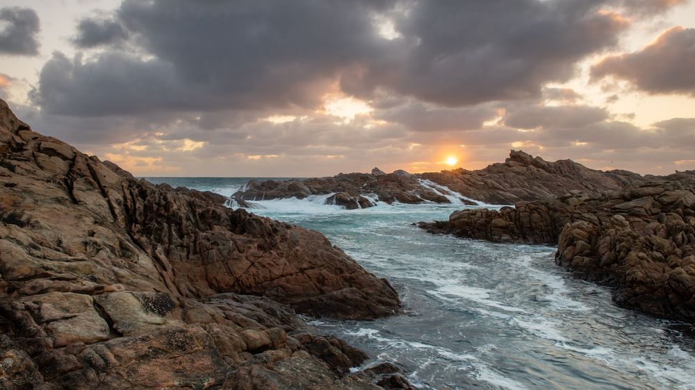 Rocky coastal landscape at sunset with dramatic clouds, crashing waves, and warm golden light over rugged stone formations.