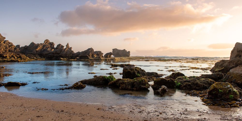 Rocky coastal beach at golden sunset with moss-covered stones, calm tidal pools, and dramatic rock formations under a glowing sky.