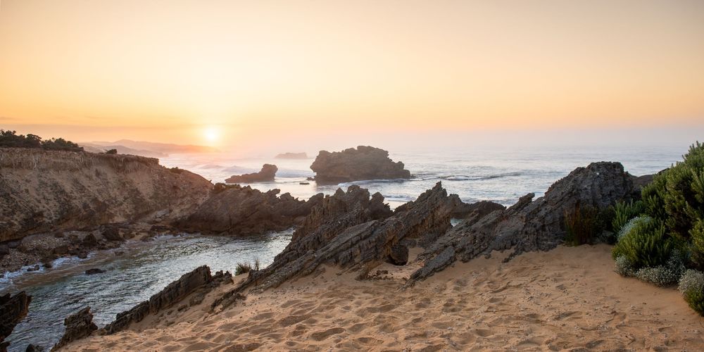 Golden sunset over rocky coastal cliffs with sandy beach and calm ocean waves in a scenic seascape.