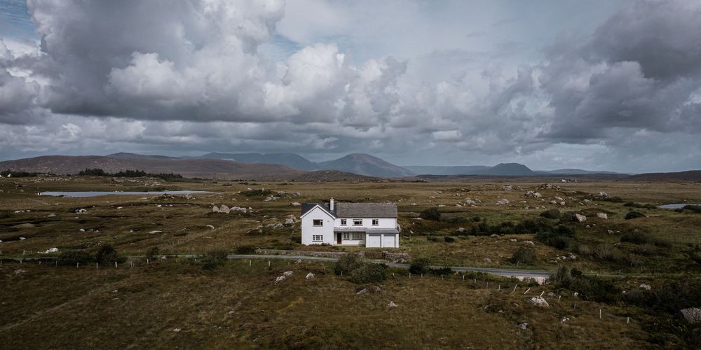 A solitary white cottage stands on a vast Irish bogland under dramatic cloudy skies with mountains in the distance.
