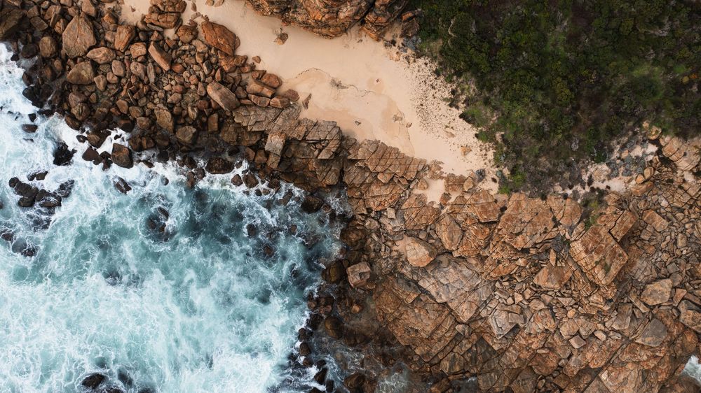 Aerial view of rocky coastline with turquoise waves crashing against brown rocks beside a small sandy beach.