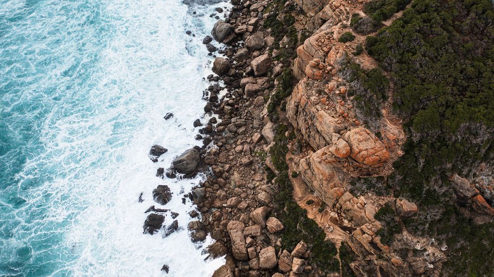Aerial view of rugged rocky coastline with turquoise ocean waves crashing against boulders and lush green cliffs.