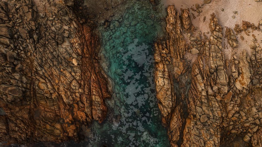 Aerial view of turquoise water flowing between rugged, textured rock formations with earthy brown and orange tones.