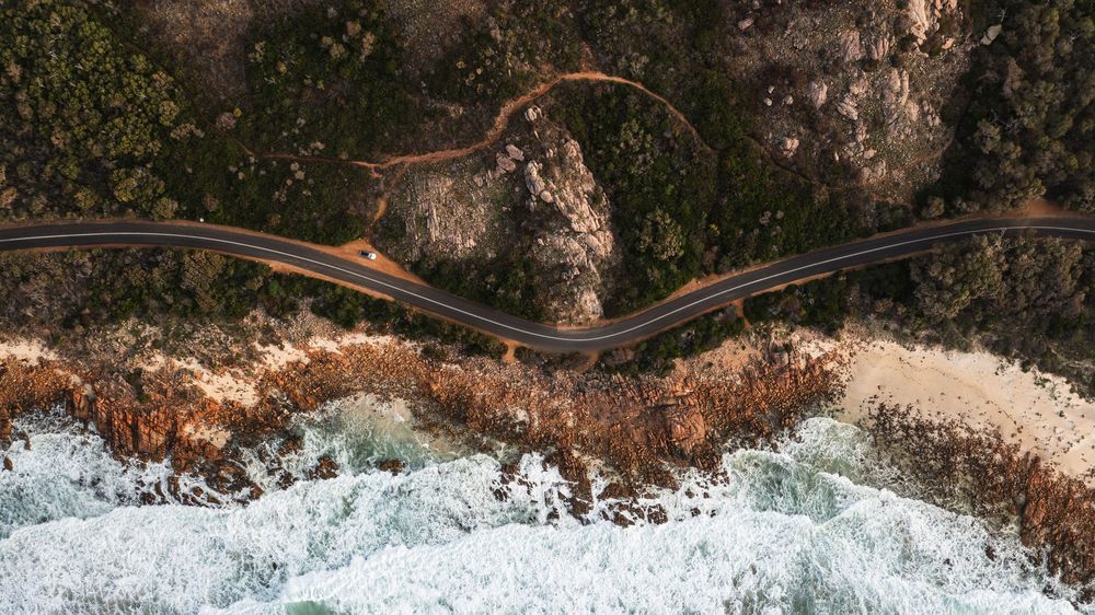 Aerial view of a winding coastal road along rocky cliffs with crashing ocean waves and lush green trees.