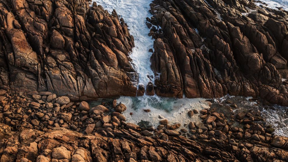 Rocky coastal cliffside with crashing ocean waves flowing through a narrow crevice surrounded by boulders and rugged stone formations.