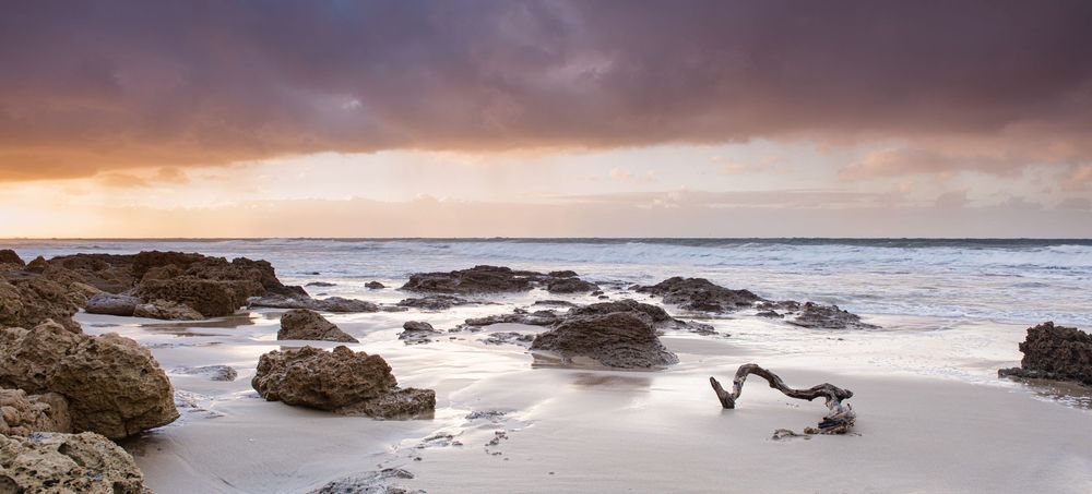 Rocky beach at sunset with dramatic cloudy sky, gentle waves washing over sand, and driftwood in the foreground.