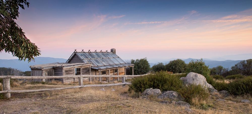 Historic rustic stone hut at sunset with panoramic mountain views, surrounded by dry grassland and scattered rocks in Australia.