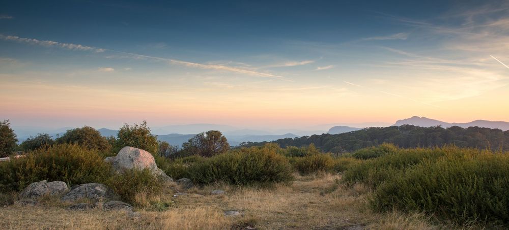 Panoramic sunset landscape with rocky terrain, shrubs, and distant mountains under a pastel sky.