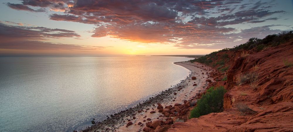 Stunning sunset over red cliffs and rocky shoreline with vibrant orange sky reflecting on calm ocean waters.
