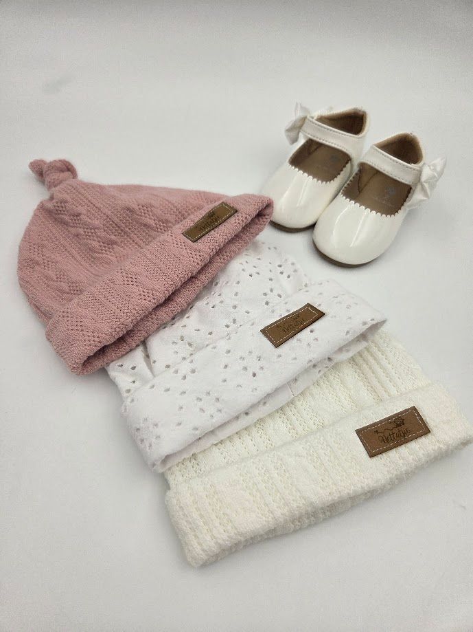 Three baby knit hats in pink, white, and cream with leather labels, paired with white patent Mary Jane shoes.