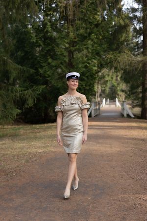 Woman in gold dress and white sailor cap walking on a tree-lined path in an outdoor park setting.