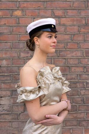 Young woman in gold ruffled off-shoulder dress and white captain's hat poses against brick wall.