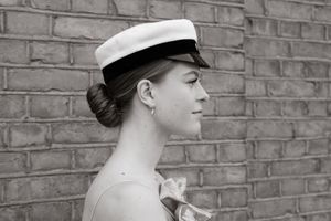 Black and white profile portrait of a young woman wearing a white sailor cap and updo hairstyle against a brick wall.