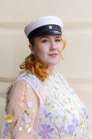 Young woman with red hair wearing a white sailor cap and floral embroidered sheer dress poses outdoors.