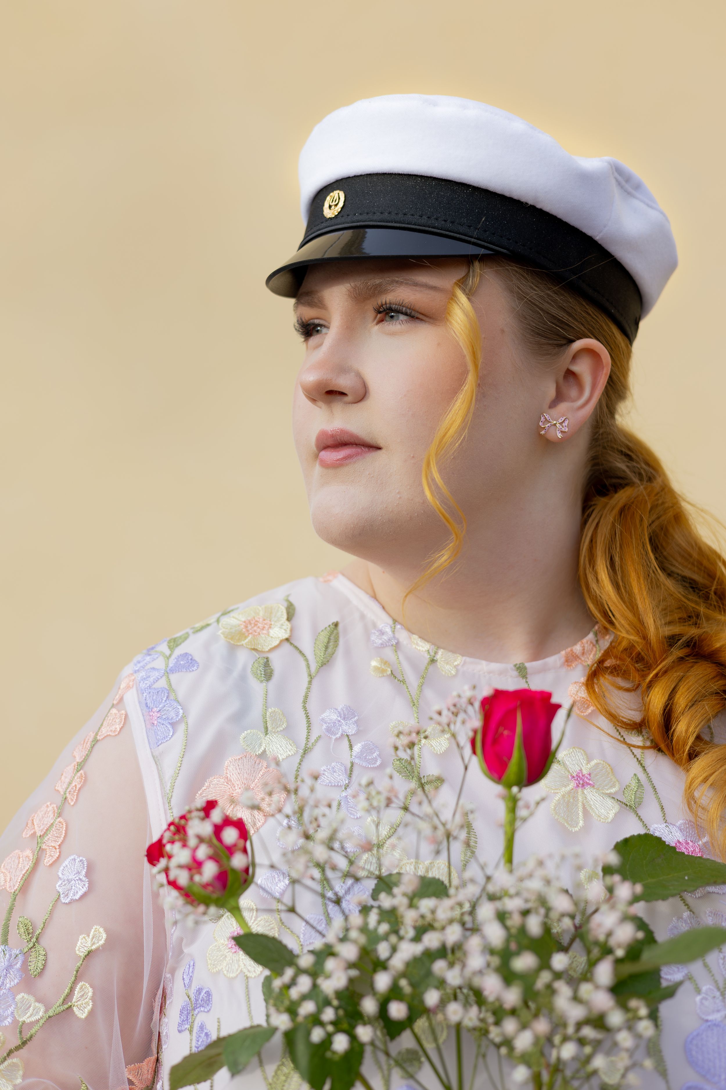 Young redhead woman in white sailor cap holding red rose and baby's breath bouquet, wearing floral embroidered dress.