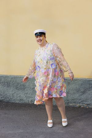 Woman in floral sheer dress and white cap smiling outdoors against yellow wall.